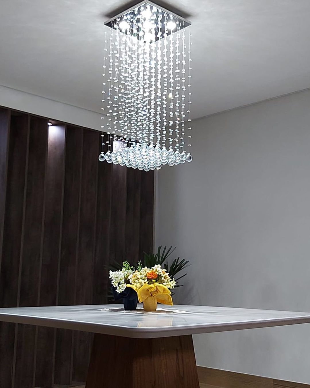 Lustre de cristal moderno pendurado no teto acima de uma mesa quadrada com um arranjo de flores Lustres Gênesis loja online de Lustres e Iluminação Decorativa